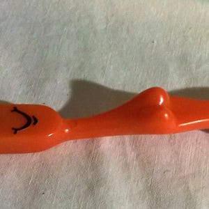 May include: An orange plastic crochet hook with a smiling face design on the handle. The hook has a long, slender metal needle extending from the face. The handle is shaped like a stylized figure.