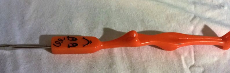May include: An orange plastic crochet hook with a smiling face design on the handle. The hook has a long, slender metal needle extending from the face. The handle is shaped like a stylized figure.