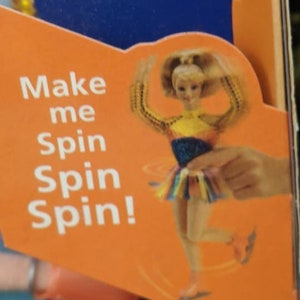 May include: An orange box featuring a doll in a skating outfit. The doll has blonde hair and is wearing a yellow, blue, and rainbow-colored outfit. Text on the box reads "Make me Spin Spin Spin!" and "Michelle Kwan Star Skater."