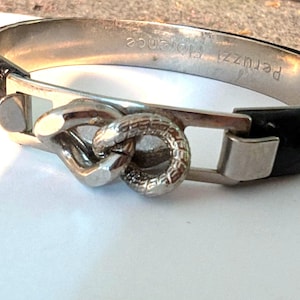May include: Silver-tone bracelet with a black leather band. The bracelet features a decorative clasp with a textured, circular design. The words "Peruzzi Florence" are engraved on the silver band.