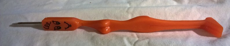 May include: Orange plastic tool with a metal tip. The tool has a stylized shape with a face drawn on the handle. The face has two eyes and a curved smile. The tool is on a white surface.