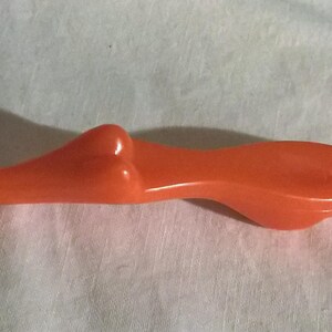 May include: Orange plastic cake decorating tool with a smiling face and curly hair design on one end. The tool has a long, slender body and a flat, angled tip. The tool is designed for decorating cakes and other baked goods.