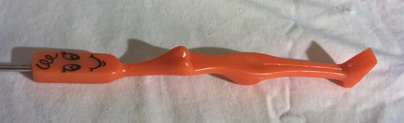 May include: Orange plastic cake decorating tool with a smiling face and curly hair design on one end. The tool has a long, slender body and a flat, angled tip. The tool is designed for decorating cakes and other baked goods.