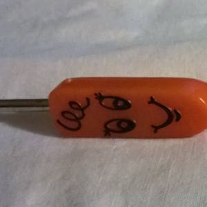 May include: An orange screwdriver with a smiling face design. The screwdriver has a flat head and a metal shaft. The handle features a cartoon face with eyes, a smile, and curly hair.