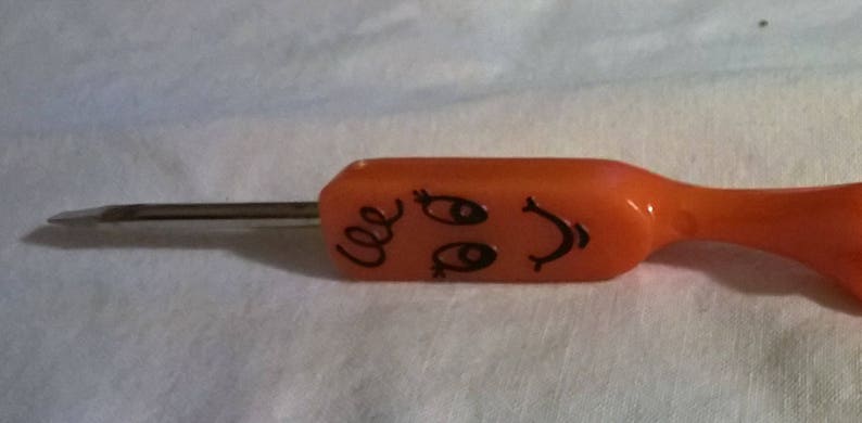 May include: An orange screwdriver with a smiling face design. The screwdriver has a flat head and a metal shaft. The handle features a cartoon face with eyes, a smile, and curly hair.