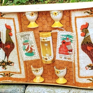 May include: A boxed kitchen set containing two white and orange rooster-patterned terry towels, four white egg cups with yellow bases and sunflower accents, and a cylindrical white and yellow timer with the text Country Style printed on a tag.