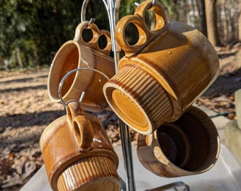 May include: Set of four brown ceramic mugs with a glazed finish hanging on a metal mug rack. The mugs have a unique design with a raised rim and a textured pattern.
