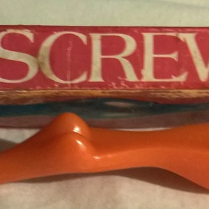 May include: An orange screwdriver with a cartoon face drawn on the handle, next to a red box with the words "SEXY SCREWDRIVER" in white. The screwdriver has a unique, curvy design.