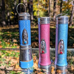 May include: Three vintage flashlights with metal casings and colored plastic bodies. The flashlights are blue, pink, and light blue. The flashlights have a silver metal band around the middle and a silver metal cap on the top.