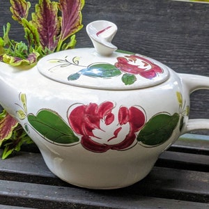 May include: White ceramic teapot with a floral design featuring red, green, and yellow flowers. The teapot has a lid with a unique swirl design.