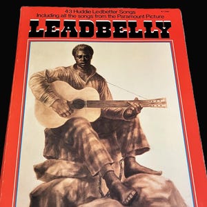 May include: Vintage album cover for Leadbelly, featuring a seated illustration of the musician playing a guitar. The cover is red with the title "LEADBELLY" in bold black letters. Text includes "43 Huddie Ledbetter Songs" and "Including all the songs from the Paramount Picture."