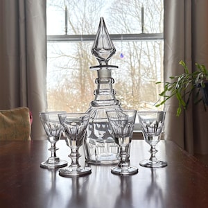 Peut inclure: Une carafe en verre transparent avec un bouchon pointu et un ensemble de cinq verres à pied assortis. La carafe et les verres sont disposés sur une table en bois sombre, avec une fenêtre et des rideaux en arrière-plan. Verrerie pour spiritueux.