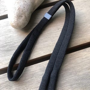 Op de afbeelding: Een zwarte, getextureerde, elastische haarband met een doorlopend lusontwerp en een klein, rechthoekig, zwart plastic verbindingsstuk dat de uiteinden van de stof bij elkaar houdt, wat zorgt voor een eenvoudig en functioneel accessoire.
