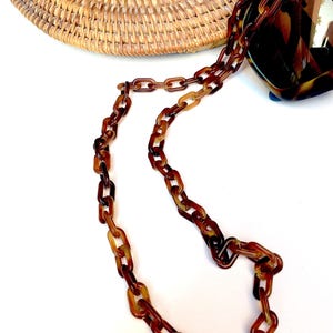 Op de afbeelding: Een bruine brilketting van acryl met schildpadpatroon, bestaande uit rechthoekige, in elkaar grijpende schakels. De ketting is bevestigd aan een bijpassende zonnebril met schildpadpatroon die gedeeltelijk op een rond, gevlochten rotan oppervlak rust.