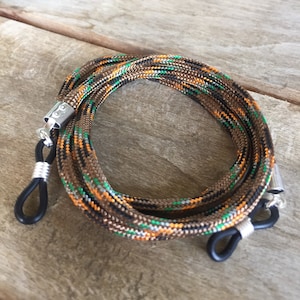 May include: A brown, black, green and orange camouflage patterned eyeglass strap with black rubber ends and silver metal clasps.