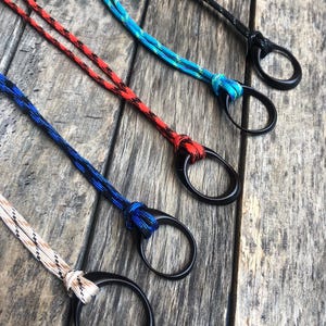 May include: A collection of five lanyards featuring black, red, blue, turquoise, and tan patterned paracord ropes. Each cord is securely knotted to a smooth, circular black ring, providing a functional attachment point for various small items.