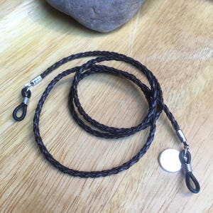 Op de afbeelding: Een bruine leren gevlochten brilketting met zilveren metalen sluitingen en een ronde zilveren hanger.