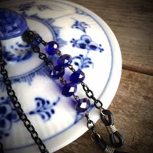Op de afbeelding: Een zwarte ketting met blauwe kristallen kralen en zwarte rubberen brillenhouders. De ketting ligt op een wit en blauw keramiek oppervlak.