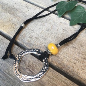 Op de afbeelding: Een ketting met een zwart koord, een grote hanger van gehamerd zilverkleurig metaal en een enkele gladde gele kraal. Het ontwerp bevat kleine zilverkleurige metalen tussenkralen langs het koord, wat zorgt voor contrast met het donkere materiaal.