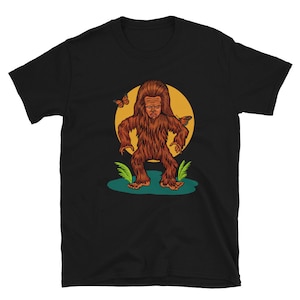May include: Black t-shirt with a graphic of a brown Bigfoot standing in a green forest with butterflies flying around it.
