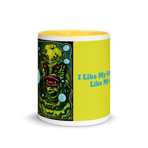 May include: A yellow and white ceramic mug with a green and blue illustration of a creature from the black lagoon. The text "I like my coffee like my monster, scary" is printed on the mug.