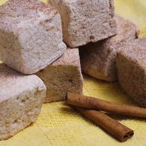 May include: A close-up of a pile of square-shaped marshmallows dusted with cinnamon, sitting on a yellow surface with two cinnamon sticks.
