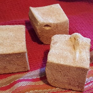 May include: Three square-shaped brown bath bombs with a grainy texture, sitting on a red and white striped towel.