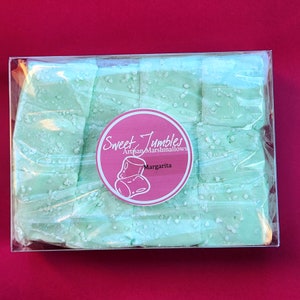 May include: A clear plastic box filled with light green square marshmallows dusted with white sugar crystals. The box has a pink circle sticker with the text "Sweet Tumbles Artisan Marshmallows" and "Margarita" in white.