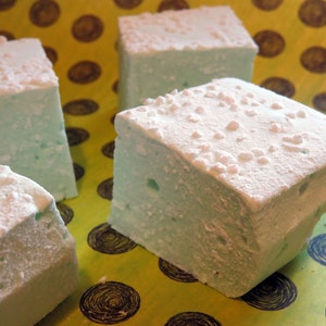 May include: Four square marshmallows, two of which are covered in white sprinkles, on a yellow and blue patterned surface.