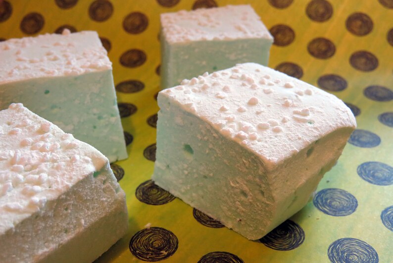 May include: Four square marshmallows, two of which are covered in white sprinkles, on a yellow and blue patterned surface.