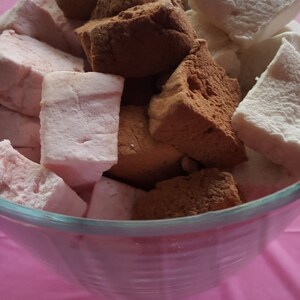 Op de afbeelding: Een glazen kom gevuld met diverse marshmallows. Sommige zijn bestrooid met cacaopoeder, andere zijn roze en sommige wit. De marshmallows zijn vierkant en liggen op een roze ondergrond.