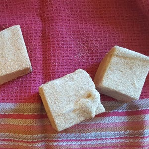 May include: Three square-shaped bath bombs on a pink and white striped towel. The bath bombs are a light brown color and have a grainy texture.