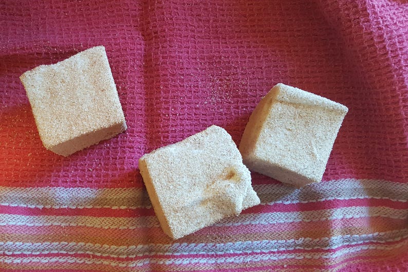 May include: Three square-shaped bath bombs on a pink and white striped towel. The bath bombs are a light brown color and have a grainy texture.