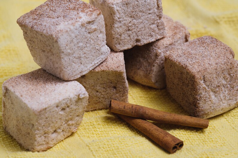May include: A close-up of a pile of square-shaped marshmallows dusted with cinnamon, sitting on a yellow surface with two cinnamon sticks.