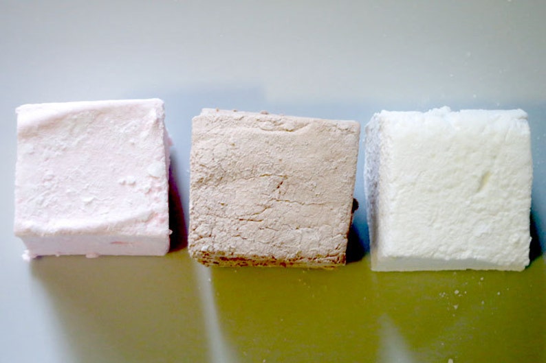 Op de afbeelding: Drie vierkante marshmallows op een rij, elk met een andere kleur en textuur. De eerste is lichtroze, de tweede is bruin en de derde is wit. De marshmallows zijn bestrooid met een fijn poeder.