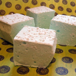 May include: Four square marshmallows, each with a light blue color and a white, powdered sugar coating, are arranged on a yellow background with a pattern of dark blue circles.