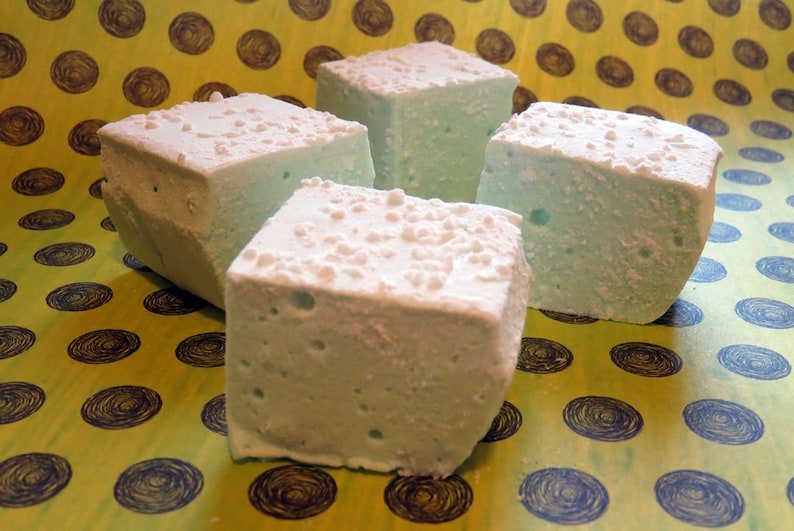 May include: Four square marshmallows, each with a light blue color and a white, powdered sugar coating, are arranged on a yellow background with a pattern of dark blue circles.