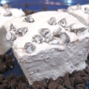 May include: Three white marshmallows covered in powdered sugar and topped with chocolate chips. The marshmallows are sitting on a blue surface with chocolate chips scattered around them.