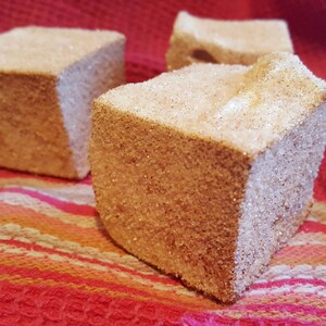 May include: Three square-shaped marshmallows covered in a layer of granulated sugar, sitting on a red and white striped fabric.