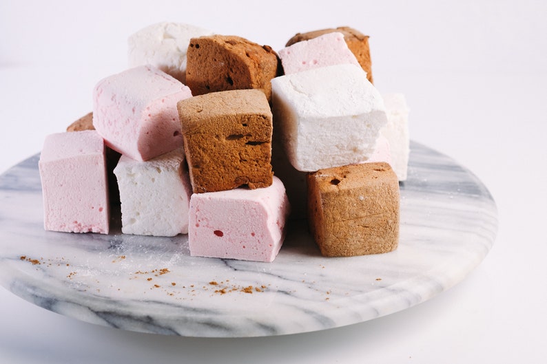 Op de afbeelding: Een stapel vierkante marshmallows in verschillende kleuren, waaronder roze, wit en bruin, gerangschikt op een marmeren serveerschaal. De marshmallows hebben een textuur. Een lichte poederlaag is zichtbaar.