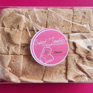 May include: A clear plastic box filled with 16 square cinnamon-flavored marshmallows. The box has a pink label that says "Sweet Jumbles Artisan Marshmallows" and "Churro".