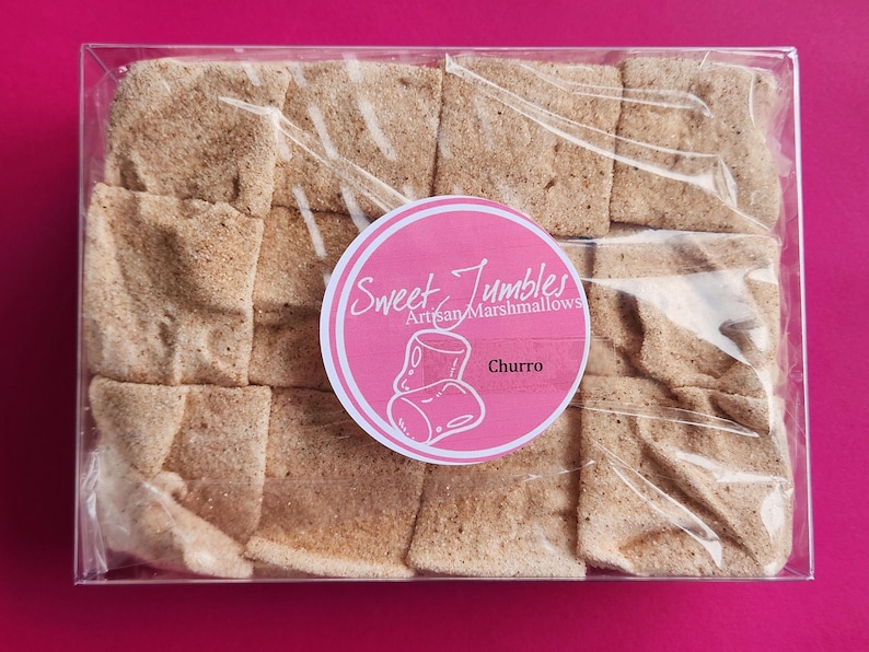 May include: A clear plastic box filled with 16 square cinnamon-flavored marshmallows. The box has a pink label that says "Sweet Jumbles Artisan Marshmallows" and "Churro".