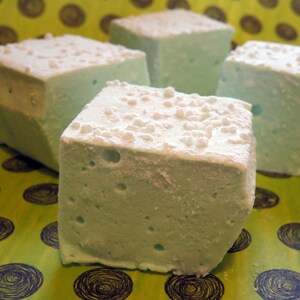 May include: Four square marshmallows, each with a light blue color and a white, grainy texture. The marshmallows are arranged on a green and brown patterned surface.