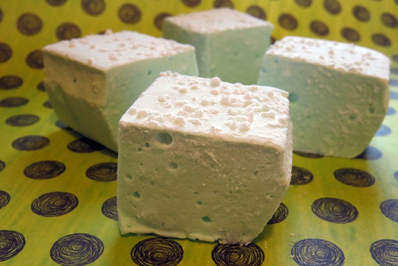 May include: Four square marshmallows, each with a light blue color and a white, grainy texture. The marshmallows are arranged on a green and brown patterned surface.