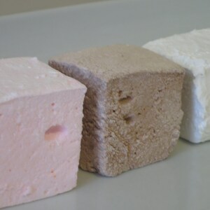 Op de afbeelding: Drie vierkante marshmallows op een rij, elk met een andere kleur. De eerste is lichtroze, de tweede is lichtbruin en de derde is wit. De marshmallows hebben een zacht, getextureerd uiterlijk.