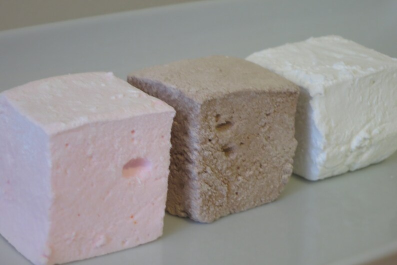 Op de afbeelding: Drie vierkante marshmallows op een rij, elk met een andere kleur. De eerste is lichtroze, de tweede is lichtbruin en de derde is wit. De marshmallows hebben een zacht, getextureerd uiterlijk.