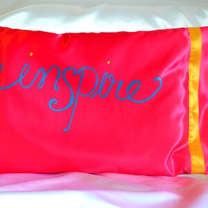 May include: A bright pink satin pillow with a yellow satin trim. The word "inspire" is written in blue cursive on the front of the pillow.