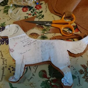 May include: A white paper pattern of a dog with handwritten instructions for sewing a stuffed animal. The pattern is laid out on a floral fabric with brown leather pieces cut out around it. The text on the pattern includes "Thread 1001", "Inner neck for Suffix spinal and (together) body", "Dyed Tolly for finished", and "Genesis Ch 3-1".