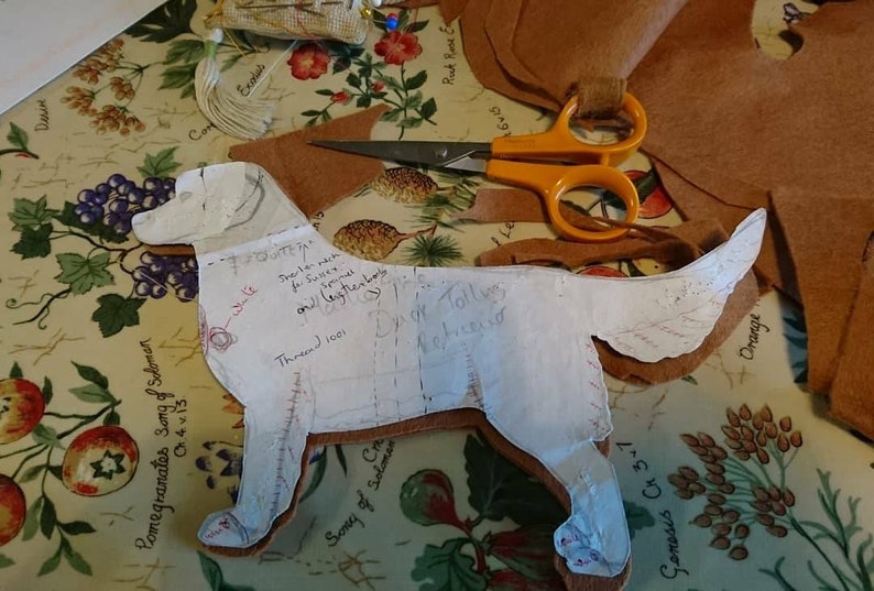 May include: A white paper pattern of a dog with handwritten instructions for sewing a stuffed animal. The pattern is laid out on a floral fabric with brown leather pieces cut out around it. The text on the pattern includes "Thread 1001", "Inner neck for Suffix spinal and (together) body", "Dyed Tolly for finished", and "Genesis Ch 3-1".