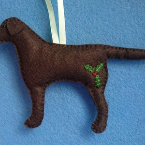 May include: A brown felt dog ornament with a holly sprig embroidered on its side. The ornament is hanging from a white ribbon.